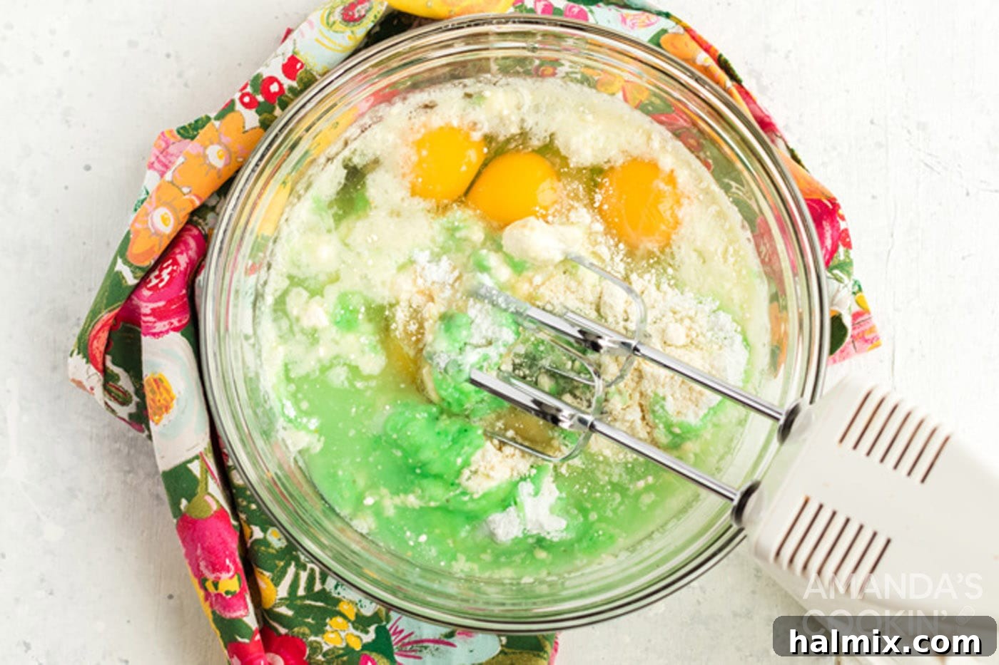 A hand mixer combining yellow cake mix, green pistachio pudding mix, water, oil, and eggs in a large mixing bowl, creating a pale green batter.