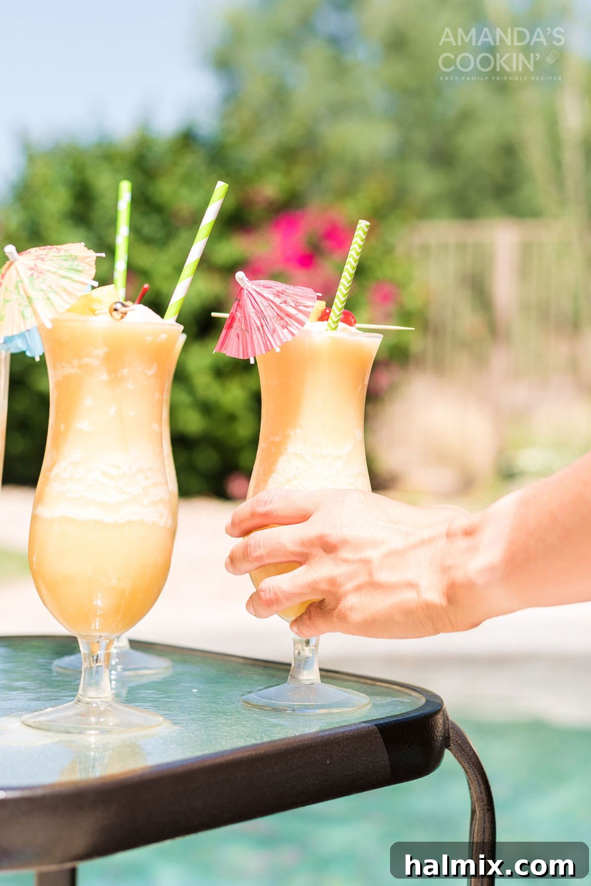 A hand reaches out to pick up a refreshing Pina Colada cocktail, glistening with condensation, from a table beside a shimmering swimming pool.