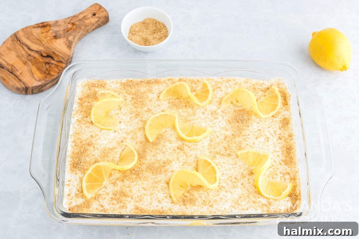 The finished lemon cream cheese pudding dessert in a pan, beautifully garnished with lemon slices and graham cracker crumbs, chilling in the refrigerator.