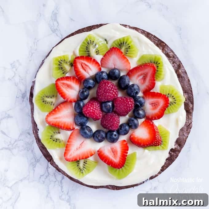An overhead shot of a vibrant brownie pizza topped with an assortment of colorful cut fruits like strawberries, kiwi, and blueberries, ready to be served.