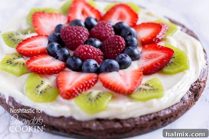 A close up of cut up fruit on top of a brownie pizza, with strawberries, kiwi, and blueberries arranged artfully.