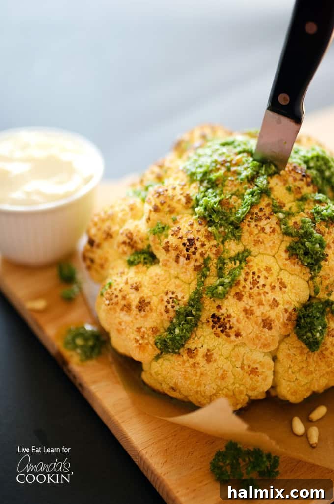 A close up of a whole roasted cauliflower topped with whipped feta and herb sauce on a wooden cutting board with a knife in the center.