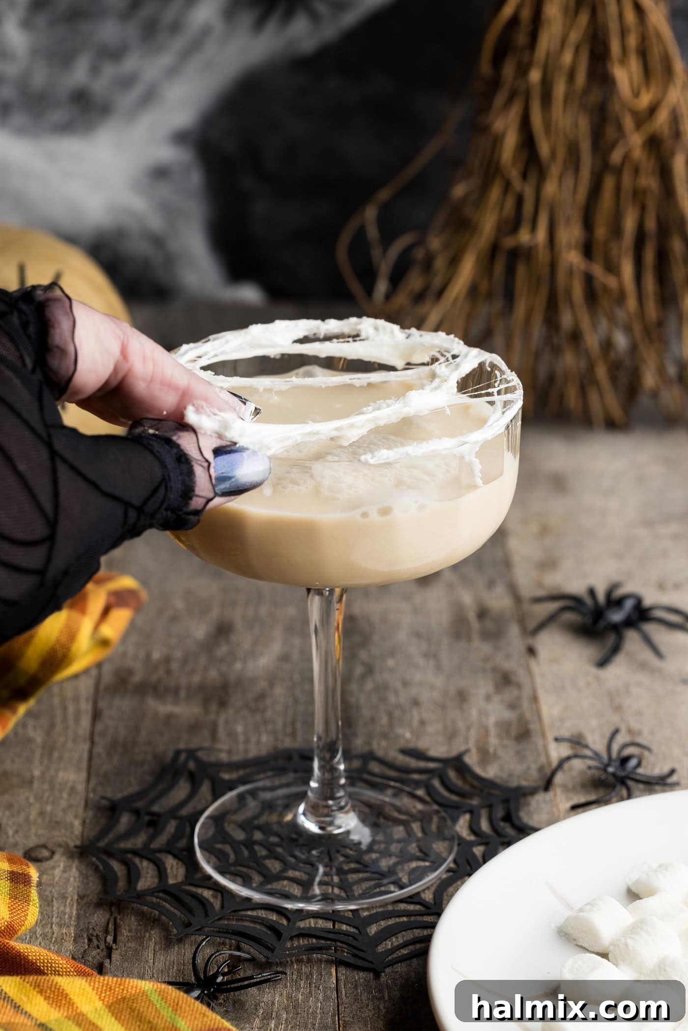 A hand stretching melted marshmallow over the rim of a cocktail glass, demonstrating the technique for creating the spiderweb topping.