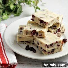 Squares of creamy White Chocolate Cranberry Fudge adorned with cranberries and pecans, presented on a pristine white plate.