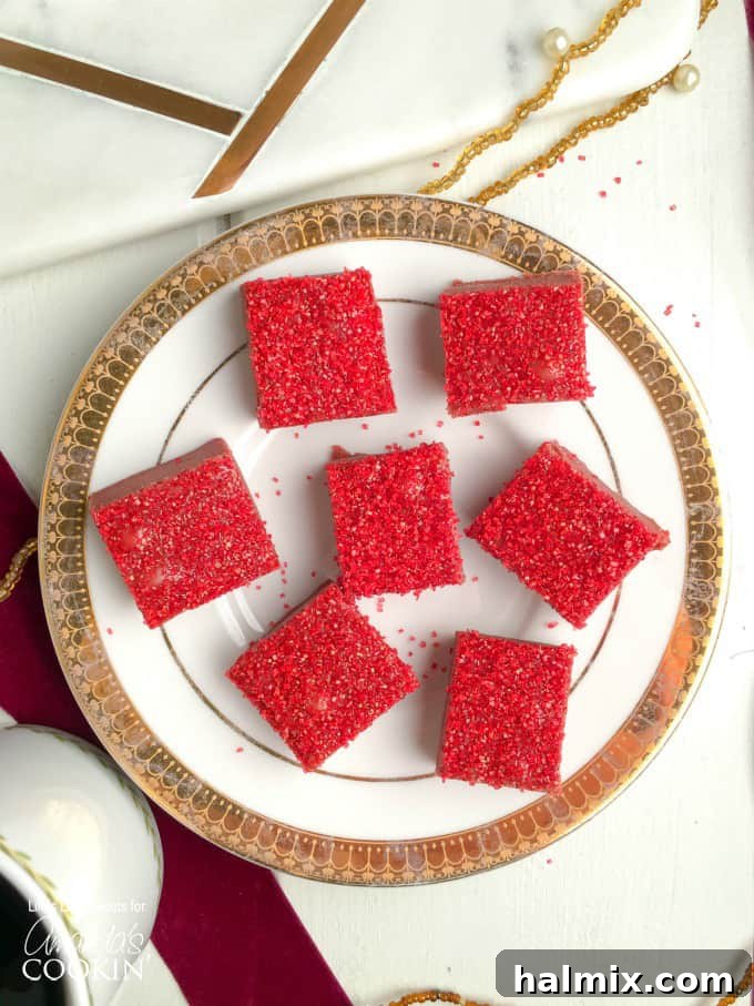 Squares of red velvet fudge with red sprinkles on top sitting on a white and gold plate.