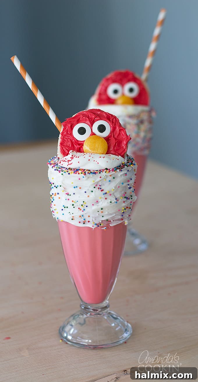 A vibrant red Elmo Freakshake in a tall glass, with a vanilla frosted rim adorned with colorful sprinkles, topped with whipped cream and an Elmo cookie.