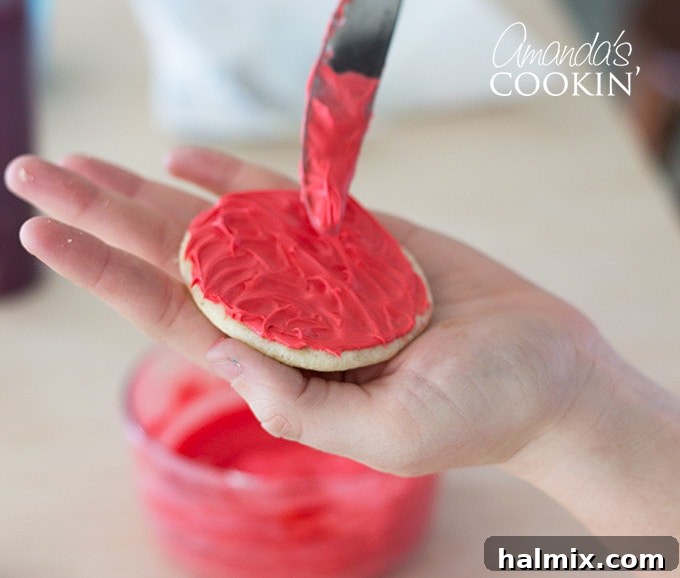 A close-up shot of vanilla frosting being spread evenly onto a sugar cookie with a butter knife, as part of the Elmo cookie decoration process.