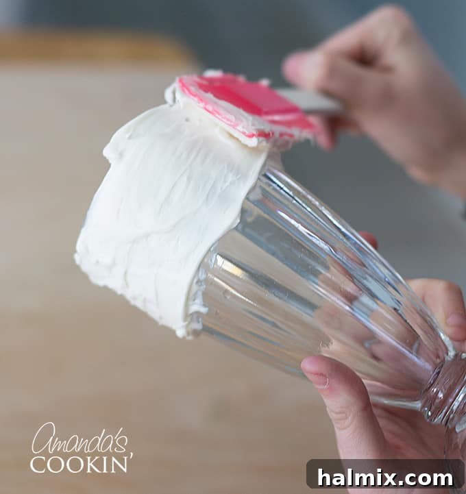 A tall milkshake glass with a thick layer of vanilla frosting being applied to its rim, a crucial step in decorating for Elmo Freakshakes.