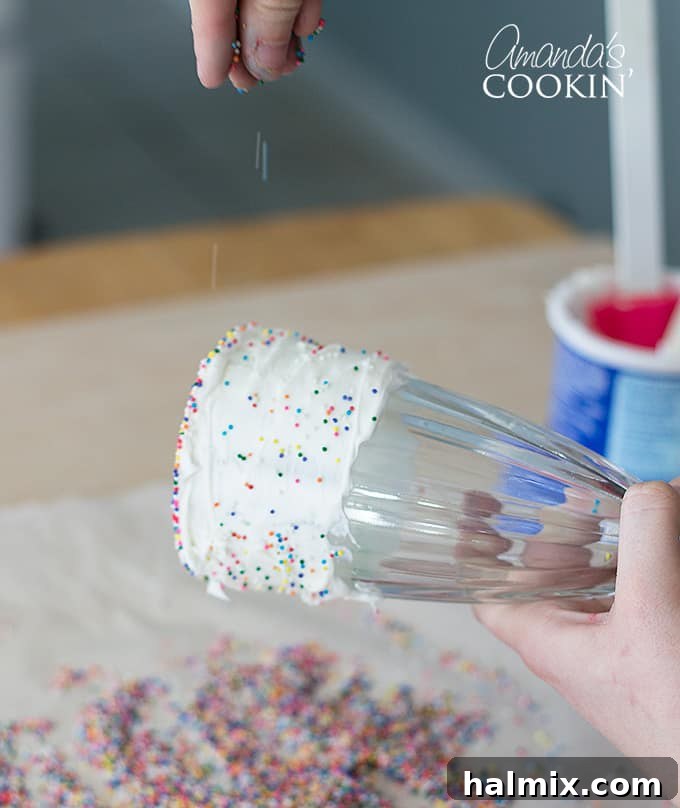 A hand sprinkling multi-colored sprinkles over the frosted rim of a milkshake glass, adding extra flair to the Elmo Freakshake decoration.