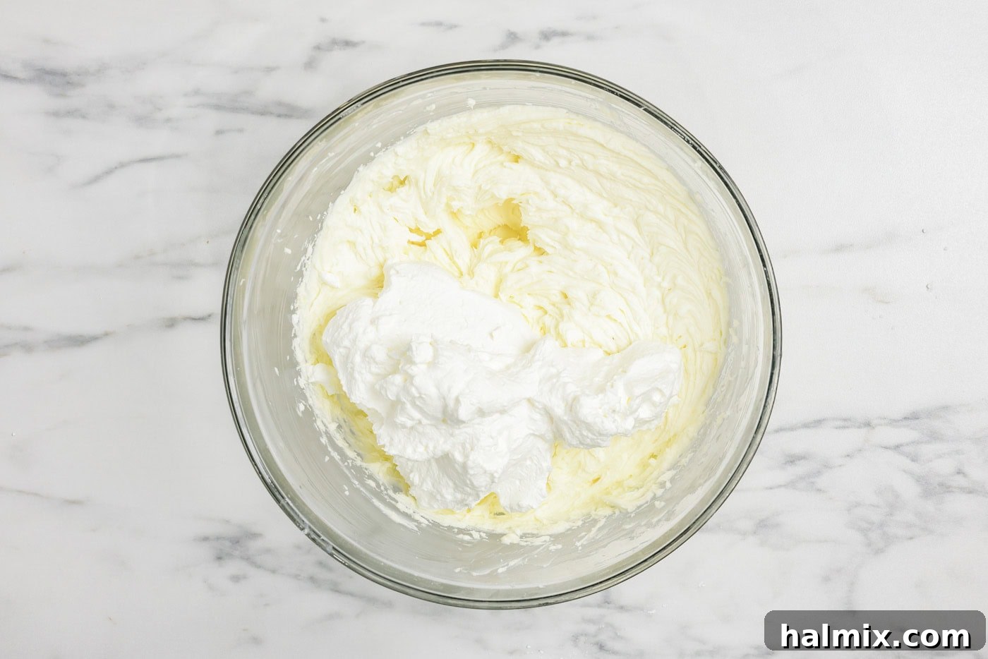 Freshly whipped cream is being gently folded into the mascarpone and cream cheese mixture in a bowl, creating a light and airy tiramisu filling.