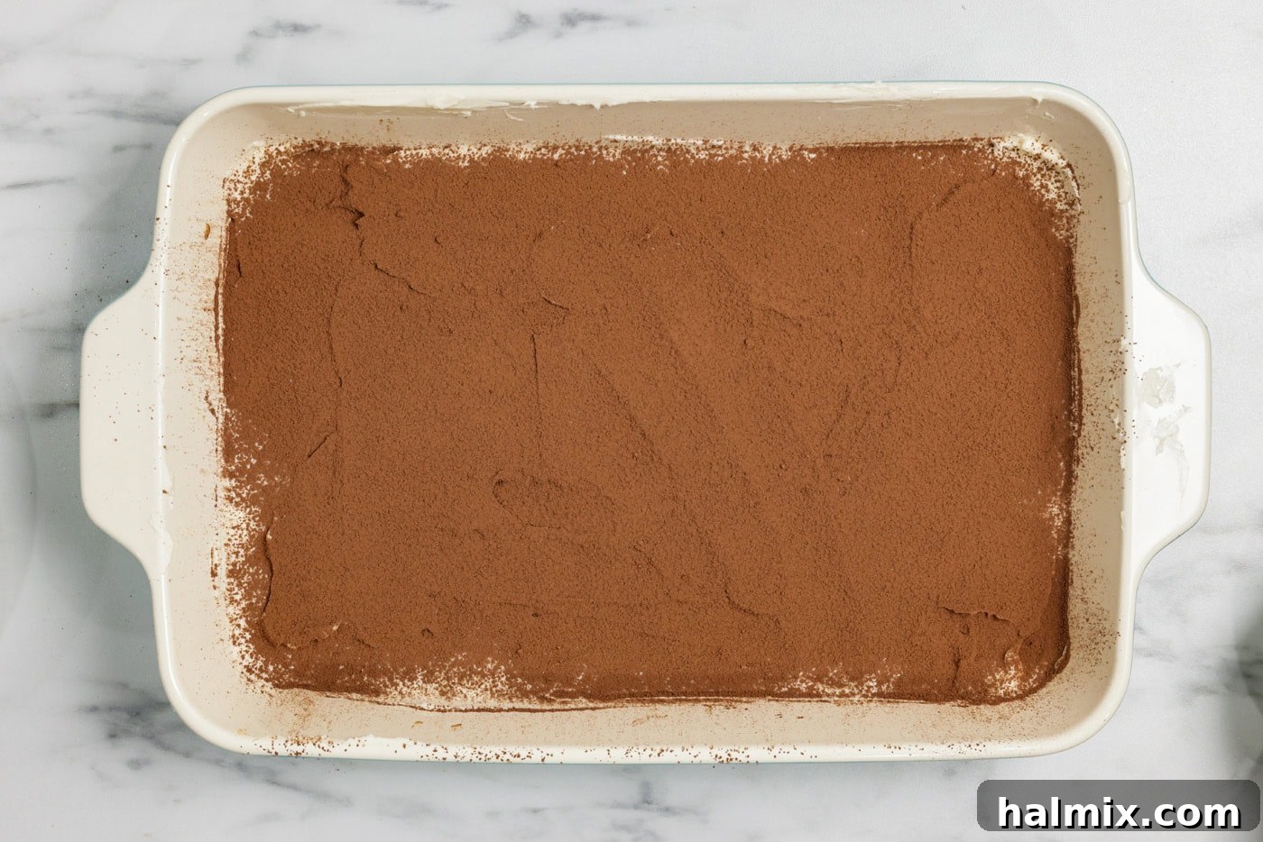 A fine layer of cocoa powder is being dusted over the mascarpone cream layer in the baking dish using a fine mesh strainer, creating a classic tiramisu look.