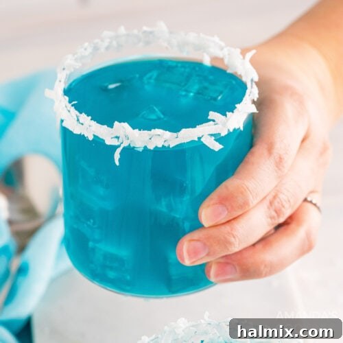 A hand holding a vibrant glass of Ocean Water Cocktail, a refreshing blue drink perfect for tropical enjoyment, set against a bright background.