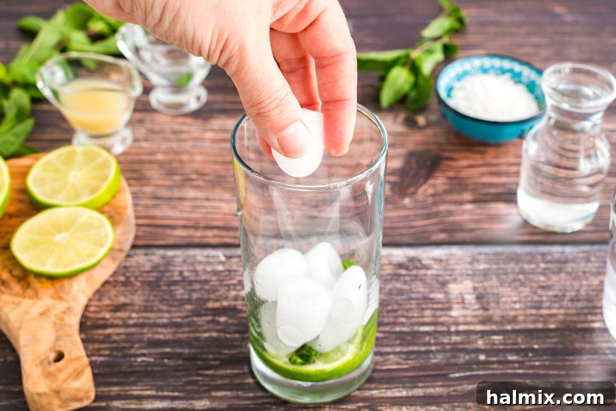 Adding ice cubes to the glass filled with muddled mint and lime, preparing for the liquid ingredients.