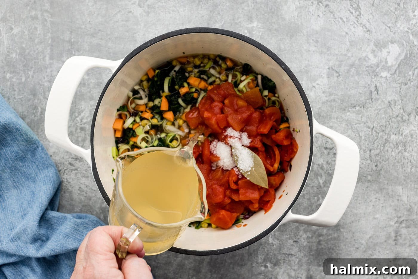 hand pouring vegetable stock into pot with diced tomatoes, bay leaf, and vegetables, preparing to boil