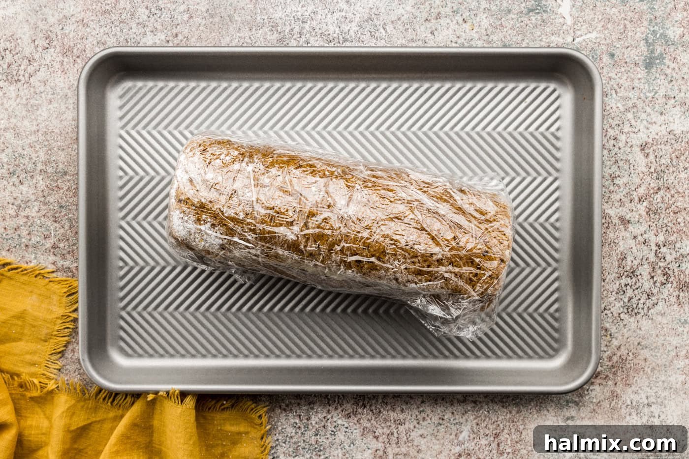 Pumpkin cake roll tightly wrapped in plastic wrap, ready to chill in the refrigerator to set the frosting.