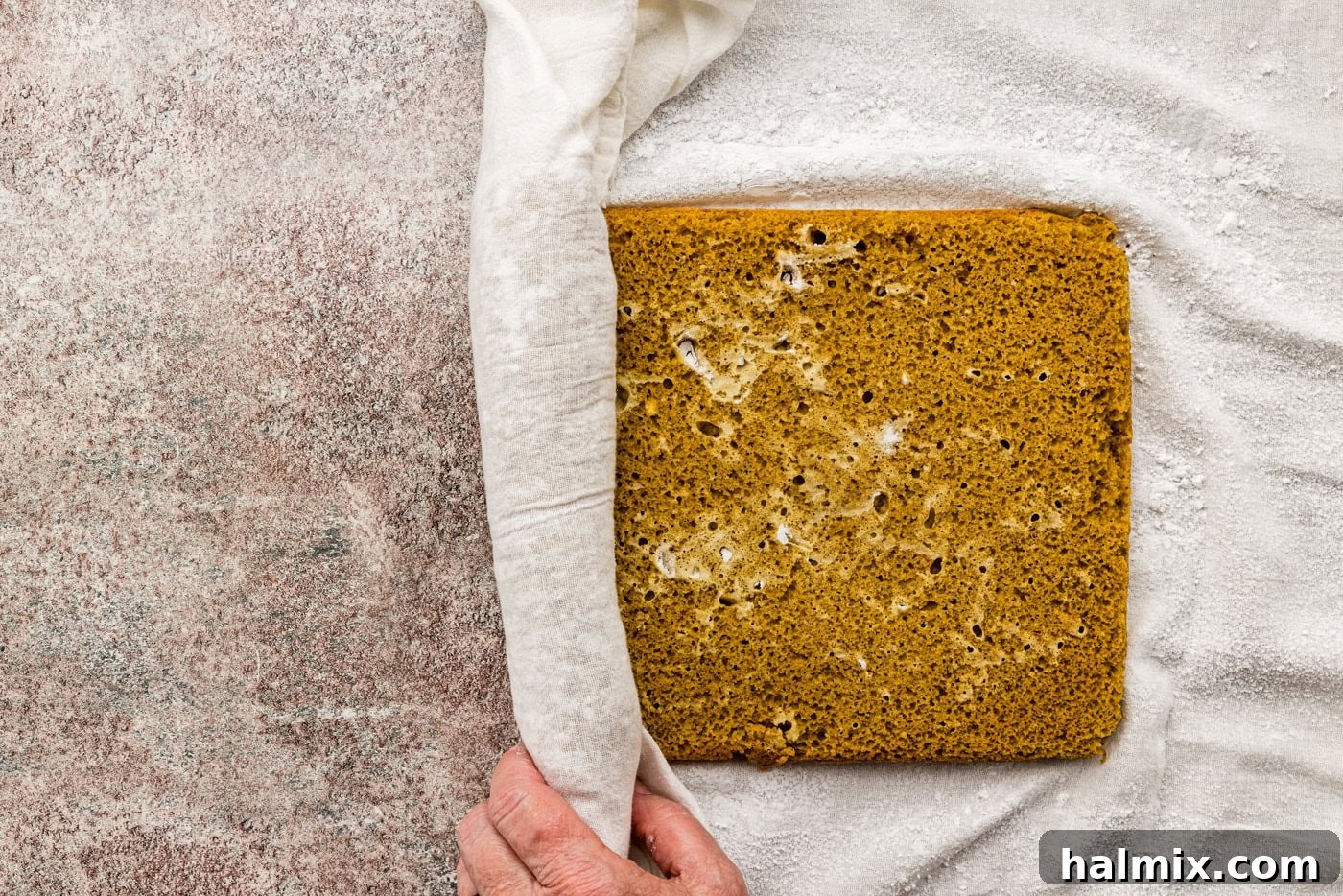 Gently rolling the warm, baked pumpkin cake into a sugar-dusted kitchen towel, a crucial step for flexibility and shape.