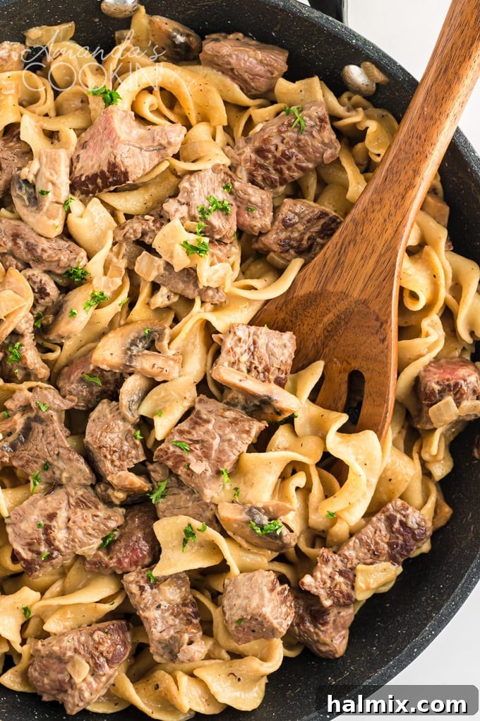 A pan filled with classic beef stroganoff, featuring tender steak slices and creamy mushroom sauce, served with a wooden spoon.