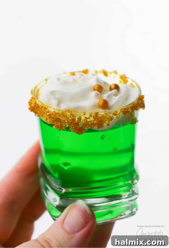 A hand holding a vibrant green jello shot, garnished with a sparkling gold sugar rim and a dollop of white whipped cream, perfect for St. Patrick's Day or a festive summer celebration.