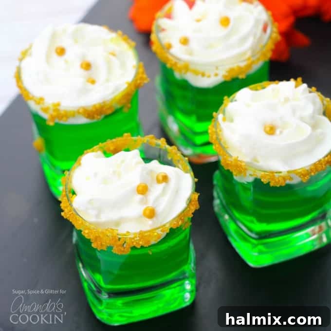 Close-up of three green jello shots arranged on a white tray, topped with whipped cream and gold pearl candies, emphasizing their festive appeal.