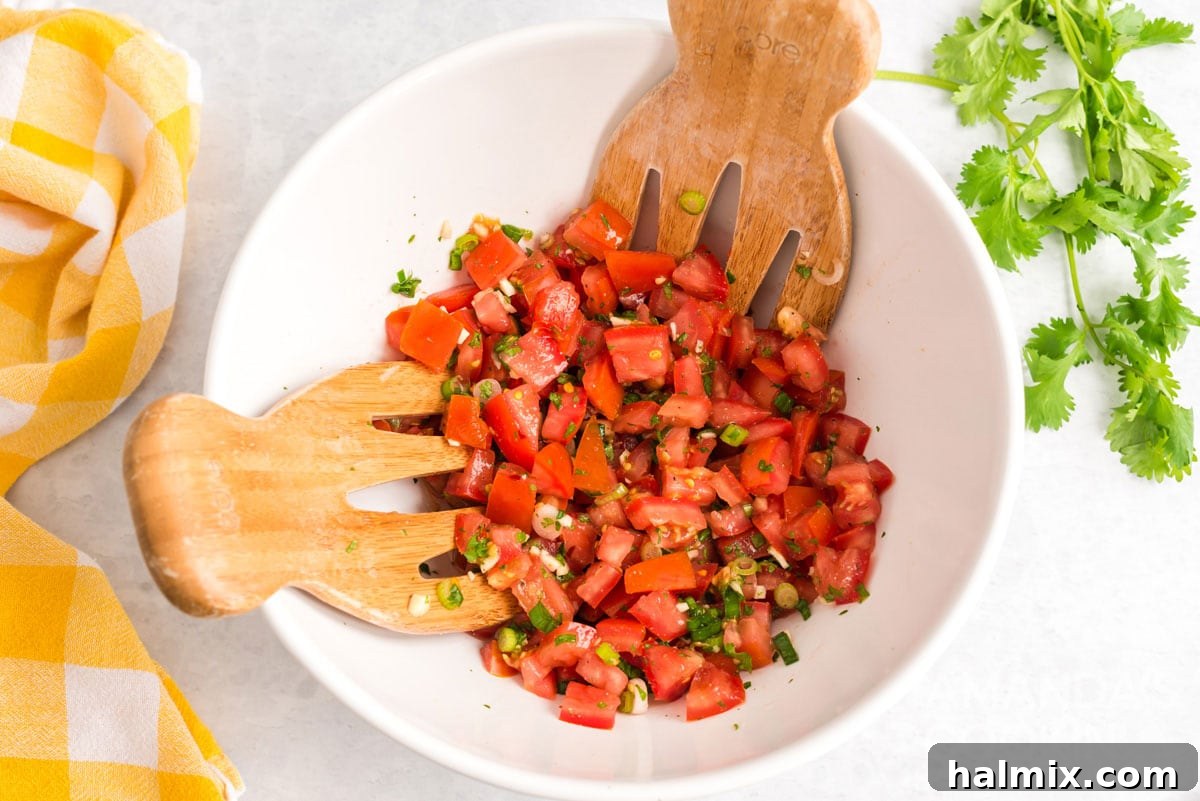 A bowl of freshly made Salsa Fresca being mixed with two salad tossing spoons, highlighting the vibrant colors and chunky texture of the salsa.