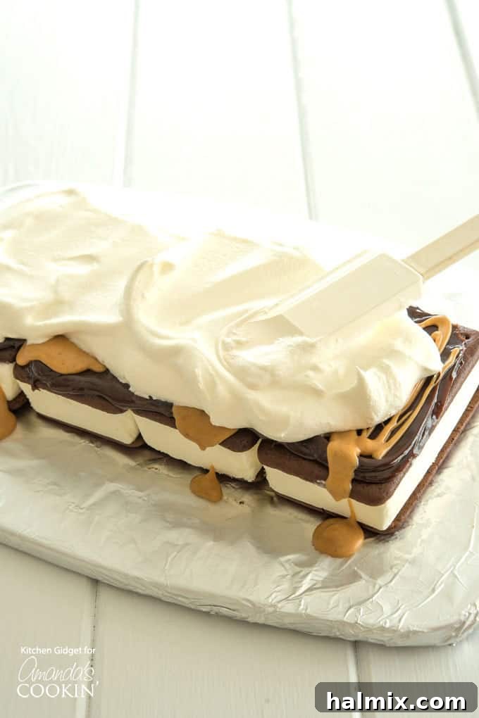 Fluffy whipped topping being carefully spread over the first layer of ice cream sandwiches, hot fudge, and peanut butter, preparing for the final topping.