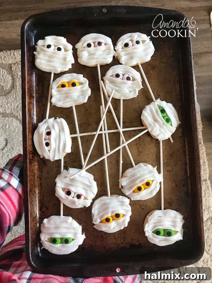 If you need Halloween food ideas for your Halloween party, be sure to check out all these sweet, savory and beverage ideas! Something for everyone. #halloweenrecipes #halloweenfoodideas #halloweenfood #halloweentreats #halloweenpartyfood #mummycookies #mozarellaeyeballs #spidertacodip #spidercookies #ricekrispypumpkins #ricekrispymonsters #dipinapumpkin #halloweenparty Close-up of a mummy cookie with candy eyes on a stick