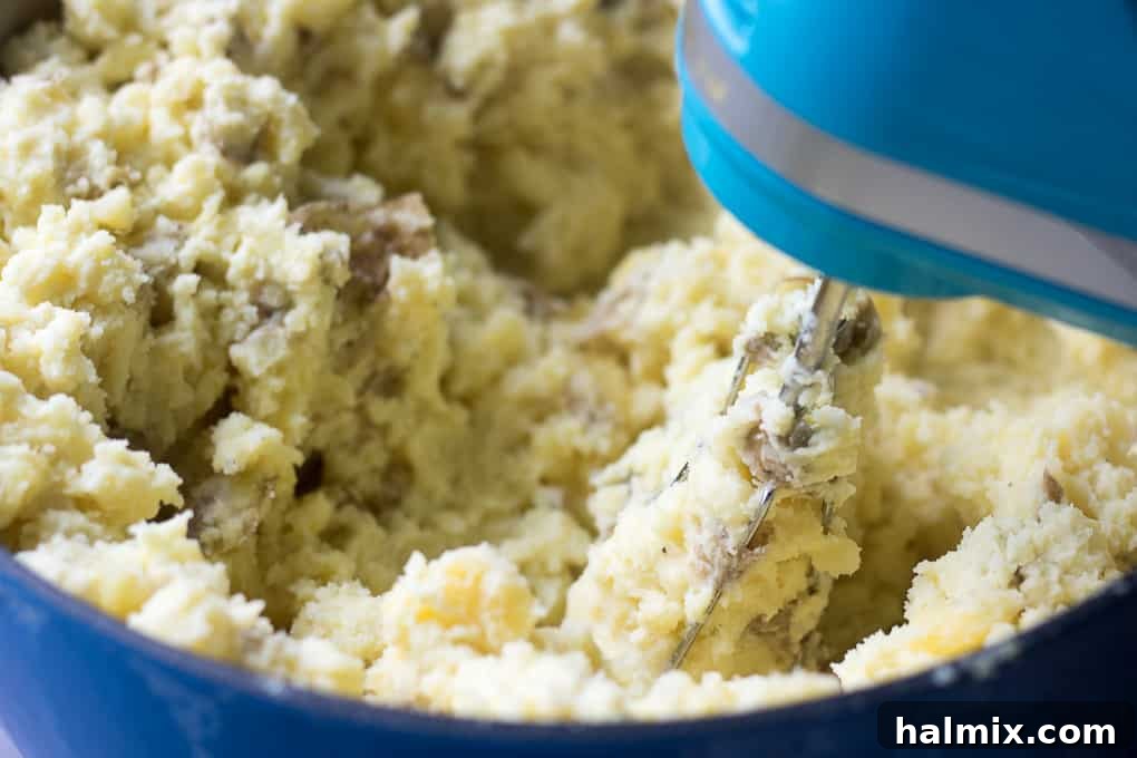 A handheld electric mixer actively mashing potatoes with butter and cream cheese.