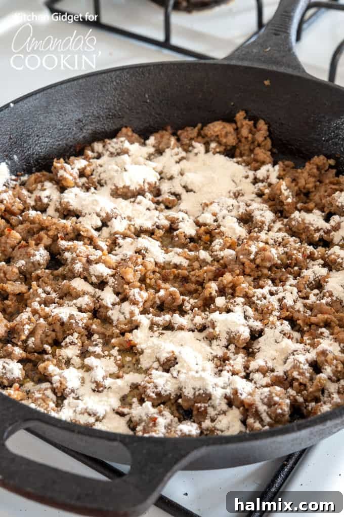 Flour being sprinkled over browned sausage and melted butter in a skillet, creating the roux for the creamy gravy.
