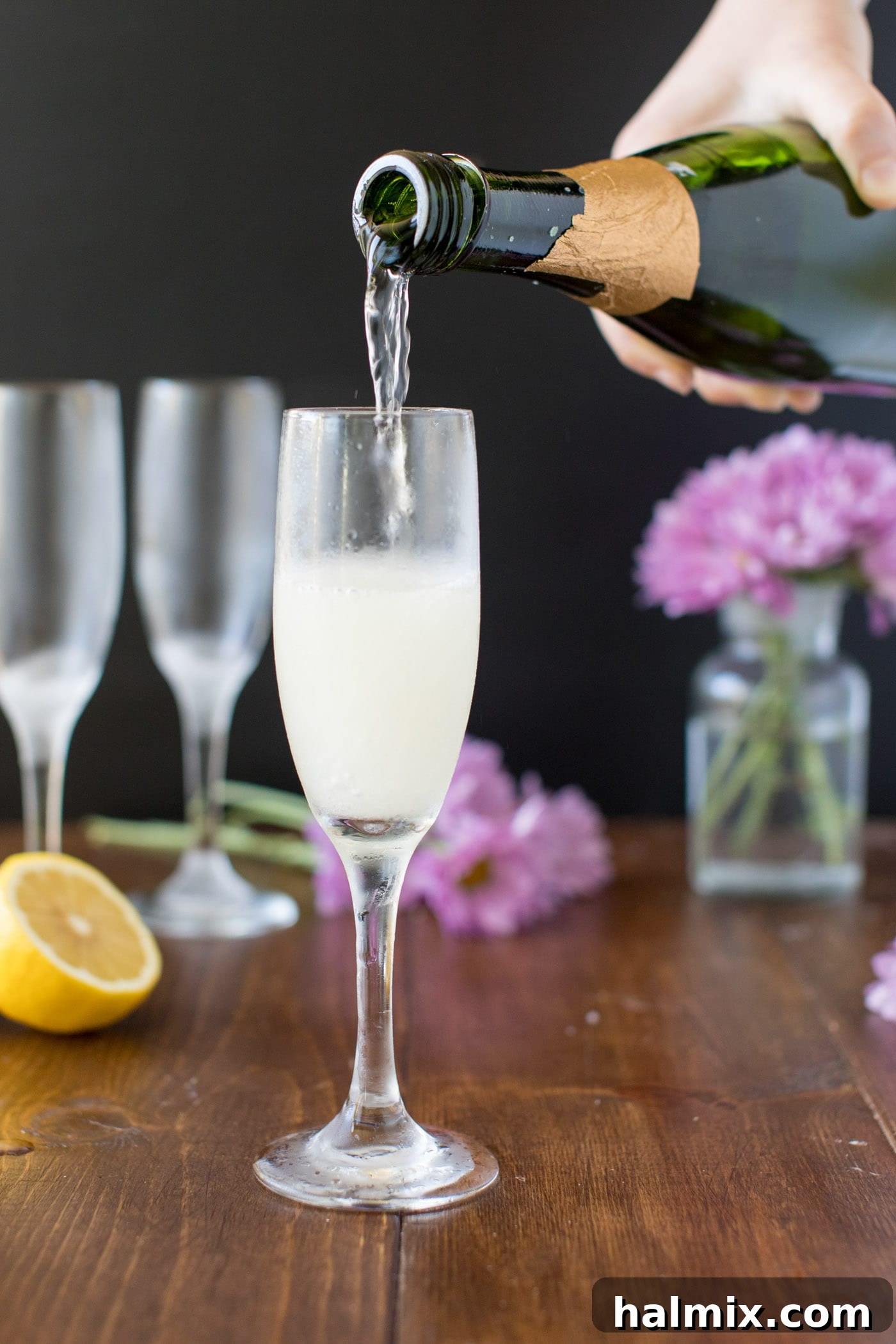 A hand gently pouring sparkling champagne into a tall, elegant flute already containing the gin and lemon mixture for a French 75.