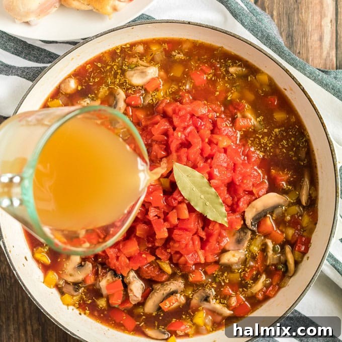 Tomatoes, broth, and bay leaf over ingredients in skillet for Chicken Cacciatore. Diced tomatoes, chicken broth, and a bay leaf added to the skillet with vegetables, forming the base of the Cacciatore stew.