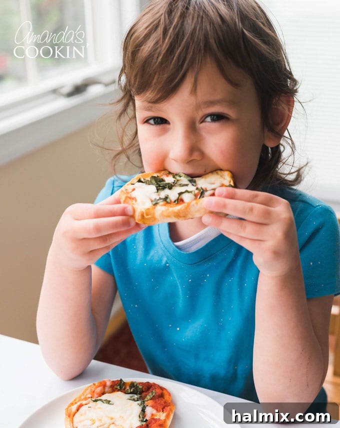 A happy little girl taking a bite from an English muffin pizza, enjoying her customized creation