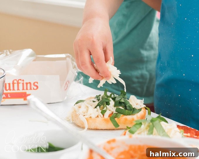 A small child carefully adding shredded cheese to their English muffin pizza, engaged in the cooking process