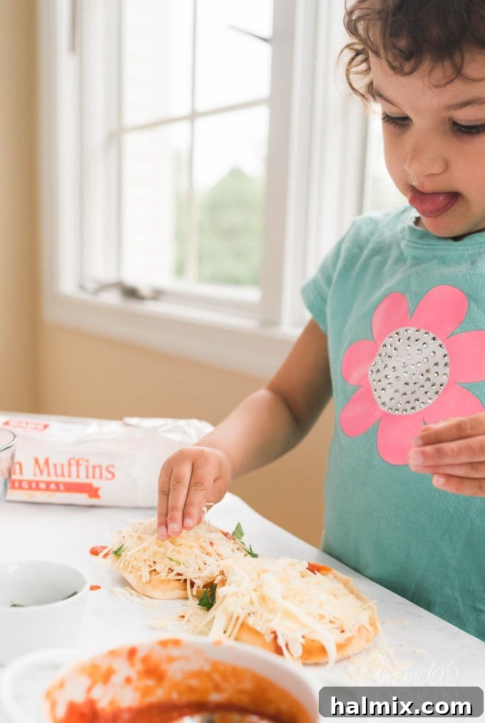 Small child focused on adding cheese to an English muffin pizza with intense concentration