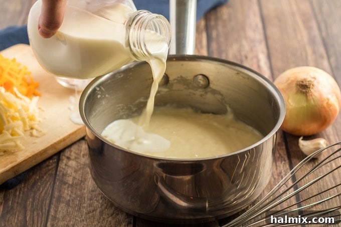 A steady stream of milk being slowly poured into a saucepan with the onion, garlic, and flour mixture, while being continuously whisked to create a smooth sauce.