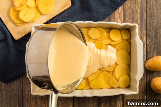 A generous pour of creamy cheese sauce being spread evenly over the first layer of thinly sliced potatoes in a baking dish.