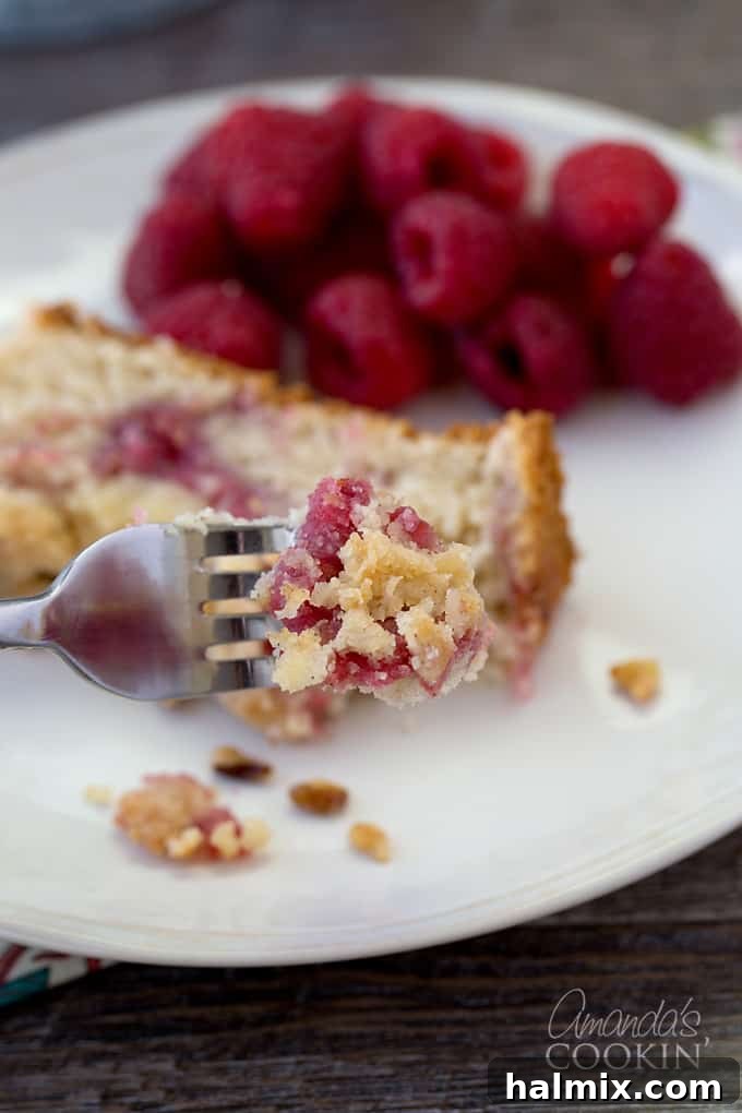 A close-up shot of a fork lifting a piece of raspberry coffee cake, with fresh raspberries scattered around and a full slice in the background.