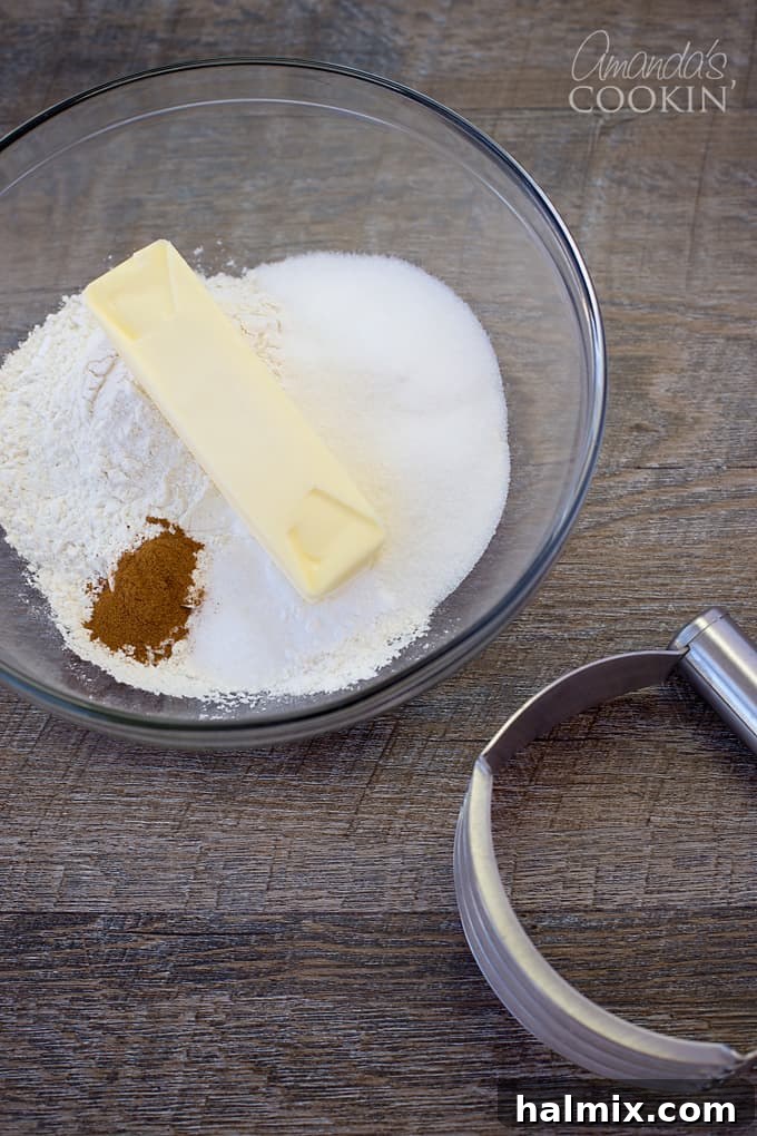 A clear glass bowl filled with dry ingredients like flour, sugar, baking powder, salt, ground cinnamon, and cold cubes of butter.