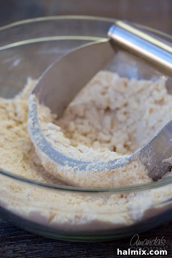 A clear glass bowl containing the coarse crumb mixture for the cake batter, with a pastry blender resting in it.