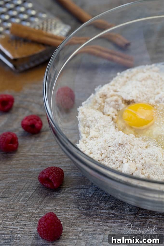 Fresh raspberries next to a clear bowl containing a flour mixture and a freshly cracked egg, ready to be incorporated into the coffee cake batter.