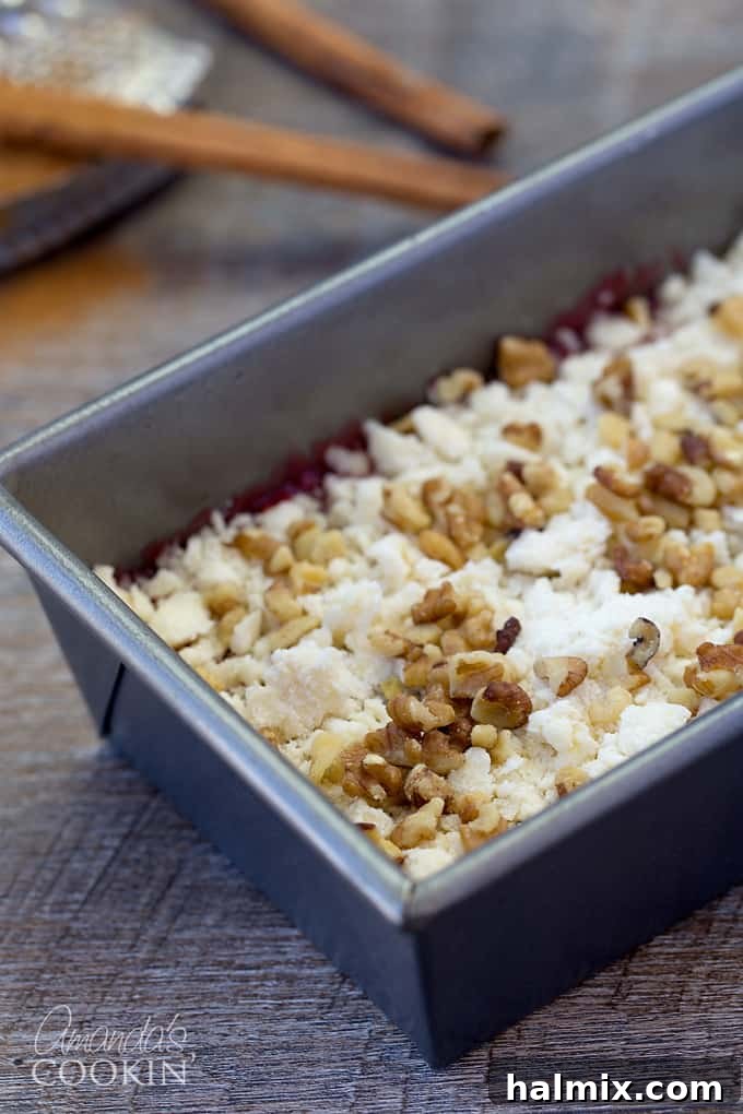 A close-up shot of a loaf pan showing the coffee cake batter, raspberry filling, crumbly streusel topping, and chopped walnuts before baking.