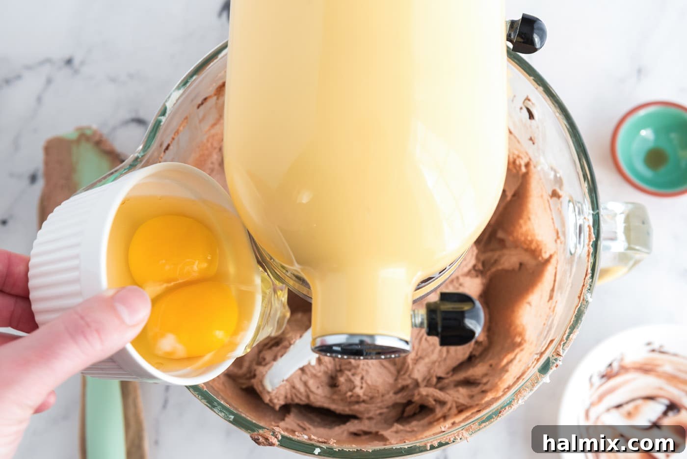 Two eggs being cracked into the chocolate mixture in a stand mixer bowl during the mixing process.