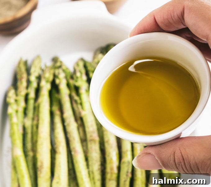 a hand holding a bottle of olive oil, drizzling it over fresh asparagus in a dish, with seasonings in the background