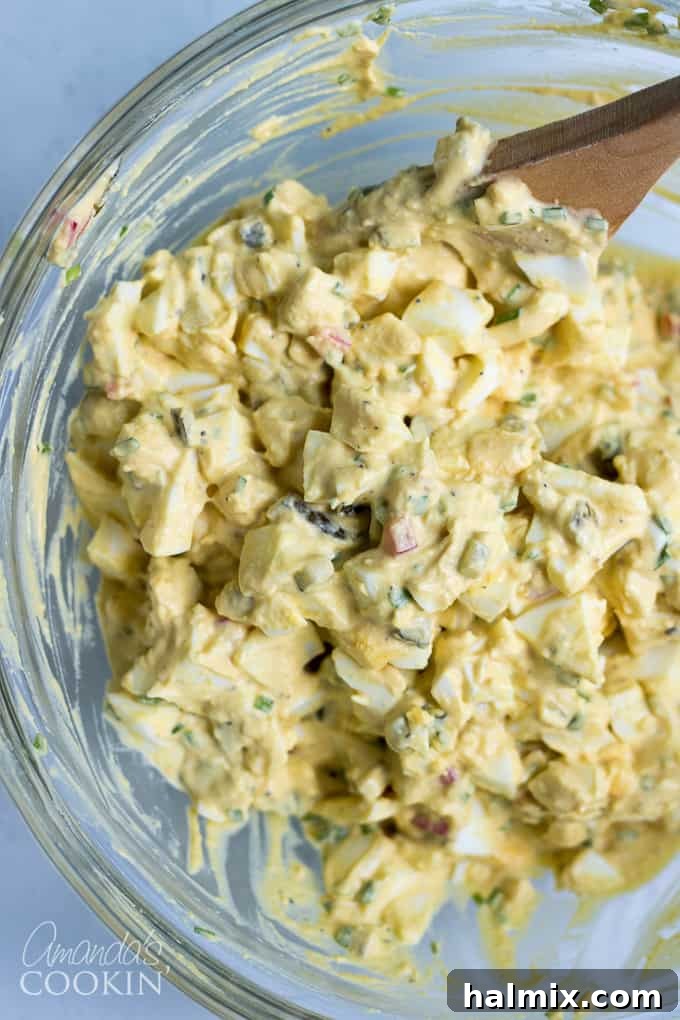Hands gently mixing all the egg salad ingredients in a bowl with a spatula, ensuring a creamy and well-combined texture before serving.