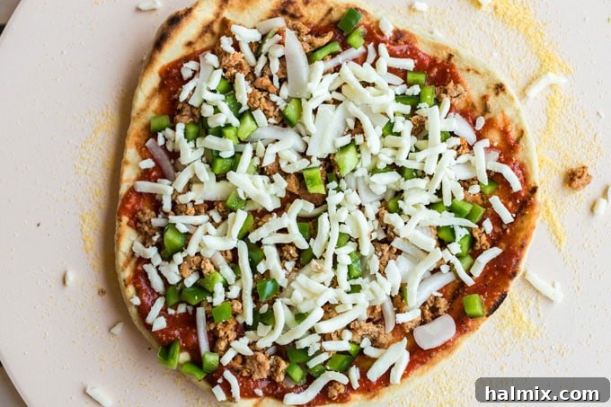 A partially cooked pizza crust fully loaded with sauce, cheese, and a variety of toppings, ready to return to the grill for final cooking.