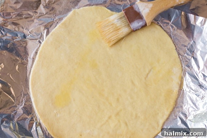 A hand using a basting brush to generously apply olive oil onto the surface of a raw pizza dough on foil.