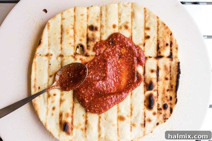A par-baked pizza crust on a cutting board, with a spoonful of red pizza sauce being spread evenly across its surface.