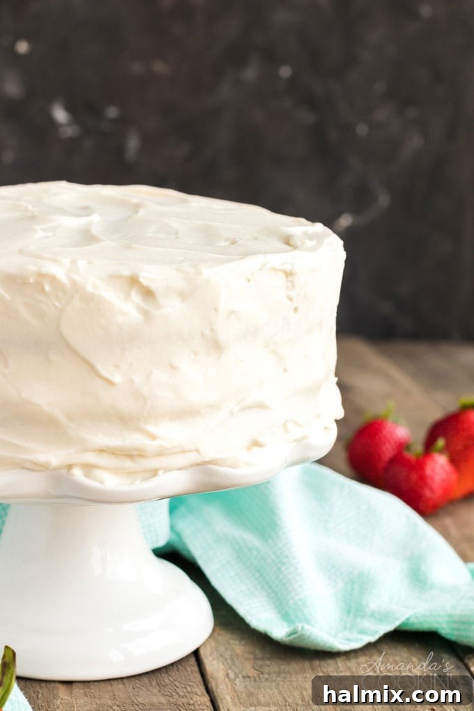 Timeless Vanilla White Cake 12 frosted white cake on a cake stand with a blue towel and strawberries in the background