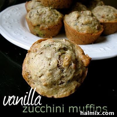 A close up photo of a vanilla zucchini muffin with a plate of them in the background, highlighting its moist texture and golden raisins.