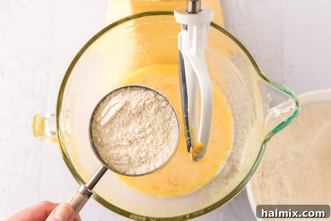 Cup of flour being added over a bowl of orange cupcake batter in a stand mixer.