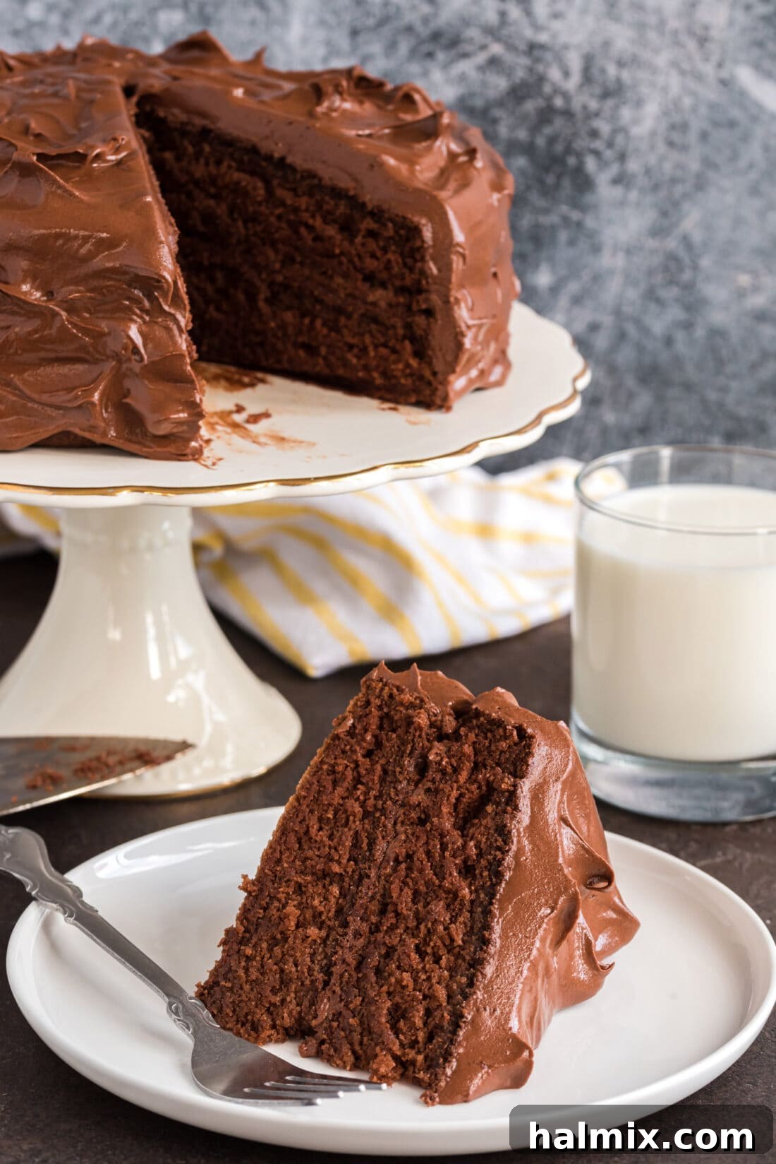 Slice of Nigella's Chocolate Fudge Cake on a plate with a fork with the rest of the cake in the background